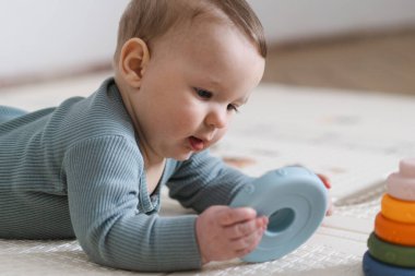 Yumuşak bir oyun hasırının üzerinde karnının üzerine uzanmış duyusal bir oyuncakla oynayan sevimli bir bebek. Bebek, oyuncağı keşfederken odaklanmış ve meraklı, erken öğrenme ve motor beceri geliştirmeyi destekliyor.