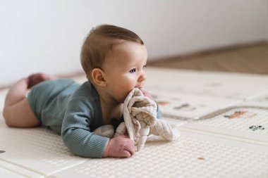 Yumuşak bir oyun hasırının üzerinde karnının üzerine uzanmış duyusal bir oyuncakla oynayan sevimli bir bebek. Bebek, oyuncağı keşfederken odaklanmış ve meraklı, erken öğrenme ve motor beceri geliştirmeyi destekliyor.