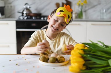 Ev mutfağında tavşan kulağı takan mutlu bir çocuk doğal boyalı paskalya yumurtalarıyla oynuyor. Sıcak aile kutlaması konsepti. Paskalya tatili, çocukluk sevinci, ev hayatı ve mevsimlik gelenek.