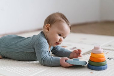 Yumuşak bir oyun hasırının üzerinde karnının üzerine uzanmış duyusal bir oyuncakla oynayan sevimli bir bebek. Bebek, oyuncağı keşfederken odaklanmış ve meraklı, erken öğrenme ve motor beceri geliştirmeyi destekliyor.