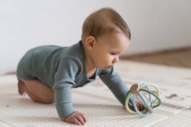 Yumuşak bir oyun hasırının üzerinde karnının üzerine uzanmış duyusal bir oyuncakla oynayan sevimli bir bebek. Bebek, oyuncağı keşfederken odaklanmış ve meraklı, erken öğrenme ve motor beceri geliştirmeyi destekliyor.