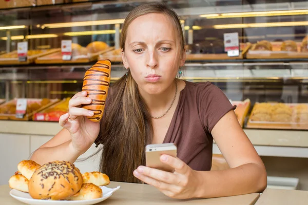 Hezká mladá žena čte zprávy na telefon a jíst chléb. — Stock fotografie
