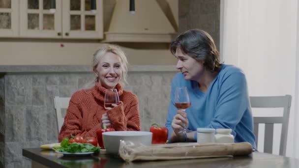 Uśmiechnięta blondynka i mężczyzna rozmawiający przy lampce wina jedzą kolację w kuchni w domu. Para rodzin pije i je domowe jedzenie na randce — Wideo stockowe