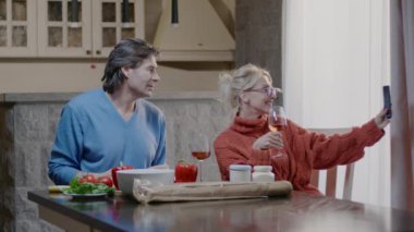 Mutlu çift telefonla selfie çekiyor ve birlikte yemek yiyorlar. Neşeli adam ve bir bardak şarapla sarışın kadın evde yemek yiyor.