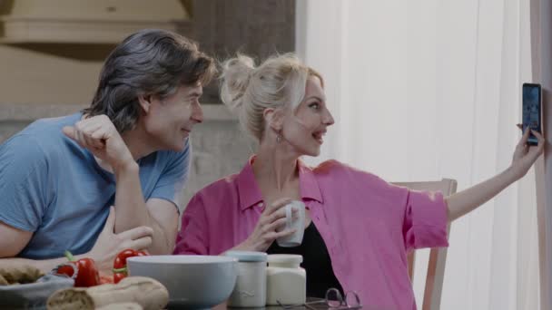Ζευγάρι κάνει selfie φωτογραφία από το τηλέφωνο και να δειπνήσετε μαζί. Χαρούμενος άντρας και ξανθιά γυναίκα με ένα ποτήρι κρασί τρώνε φαγητό στο σπίτι — Αρχείο Βίντεο