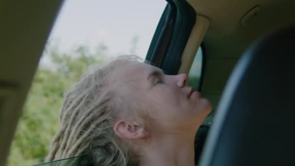 Młoda kobieta z dredami i zamkniętymi oczami podróżująca samochodem podczas podróży. Otwiera okno, aby oddychać świeżym powietrzem, porusza ręką w wiatr. Koncepcja wolności. — Wideo stockowe