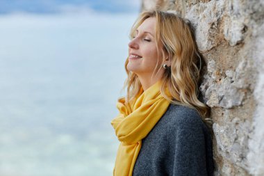 Sakin sarışın kadın gözleri kapalı sarı atkısı ve rüzgarda esen saçları denizin kenarında duruyor.