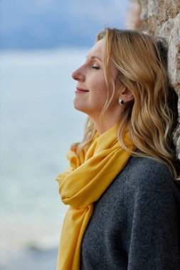 Sakin sarışın kadın gözleri kapalı sarı atkısı ve rüzgarda esen saçları denizin kenarında duruyor.