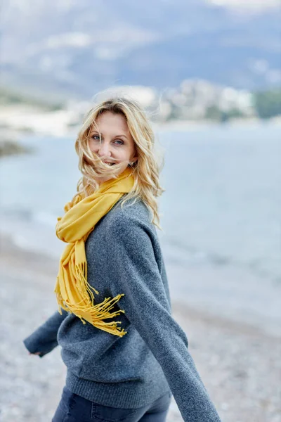 Mulher loira feliz com cachecol amarelo e seu cabelo soprando na brisa fica perto do mar Imagens De Bancos De Imagens Sem Royalties