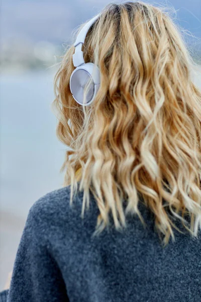 Baksidan av blond kvinna bär hörlurar lyssna positiv musik podcast från smartphone ansökan mot havet Royaltyfria Stockfoton