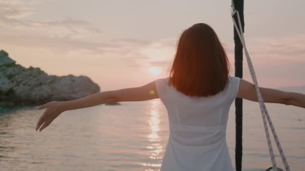 Вид сзади женщины в белом платье стоит на яхте, поднимая руки и смотрит на закат — стоковое видео