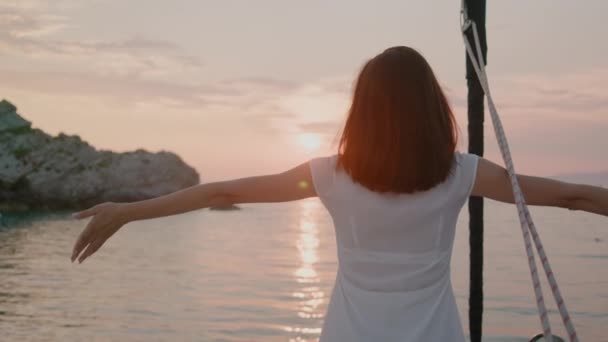 Vista posterior de la mujer en vestido blanco se para en un yate levantando las manos y mira el atardecer — Vídeos de Stock