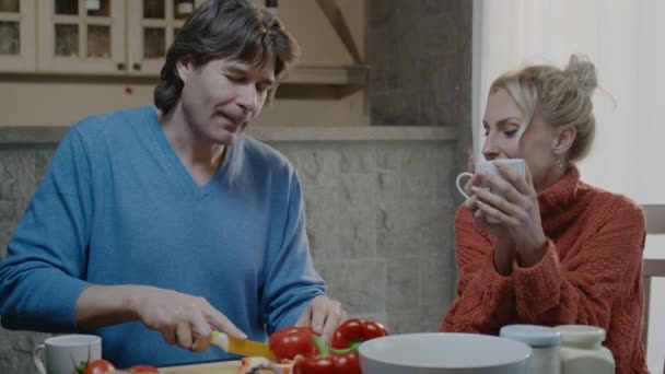 Щаслива активна сімейна пара готує їжу і сміється вдома. Красивий чоловік і блондинка вечеряють з вином — стокове відео