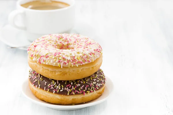 Geglazuurde donuts op een plaat en kopje koffie op witte tafel — Stockfoto