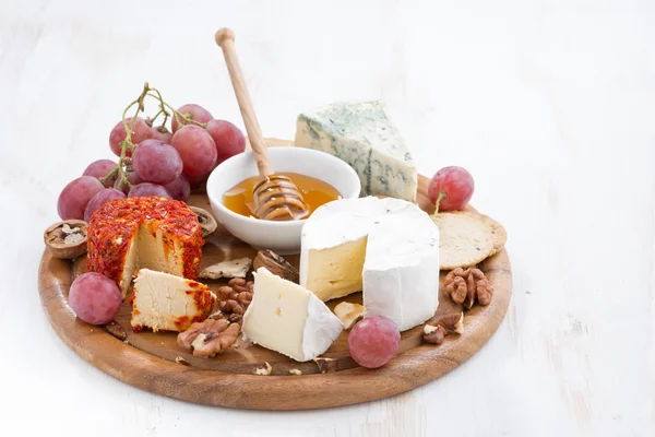 Cheeses and snacks on a board on white wooden table — Stockfoto