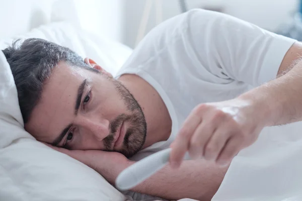 Hombre sintiéndose mal acostado en la cama y mirando el termómetro —  Fotos de Stock