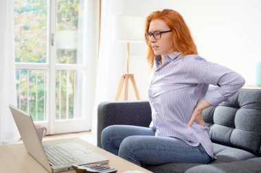 One young redhead woman suffers back pain cramp working at home clipart