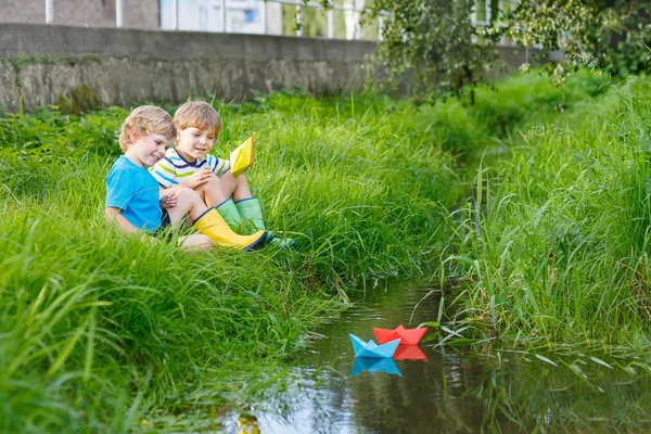 Two little brothers playing with paper boats by a river — Stock Photo, Image