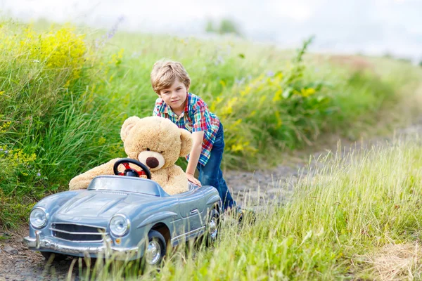 Niño pequeño conduciendo un coche de juguete grande con un oso, al aire libre . — Foto de Stock