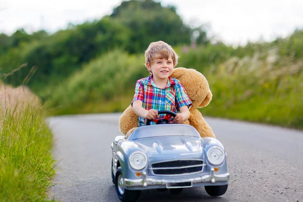 Kleiner Junge fährt großes Spielzeugauto mit einem Bären im Freien. — Stockfoto