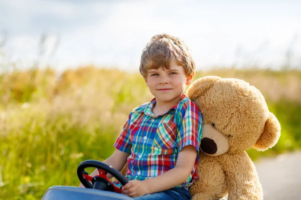 Kleine jongen jongen rijden grote speelgoedauto met een beer, buitenshuis. — Stockfoto