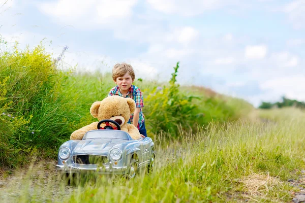Menino menino dirigindo carro de brinquedo grande com um urso, ao ar livre . — Fotografia de Stock