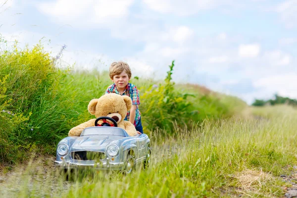 Kleiner Junge fährt großes Spielzeugauto mit einem Bären im Freien. — Stockfoto