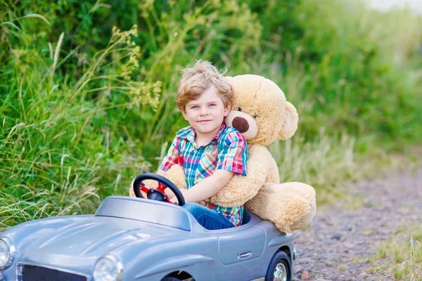 Kleiner Junge fährt großes Spielzeugauto mit einem Bären im Freien. — Stockfoto