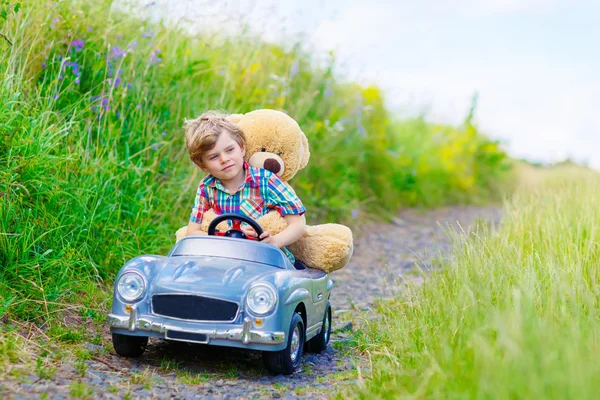 Niño pequeño conduciendo un coche de juguete grande con un oso, al aire libre . — Foto de Stock