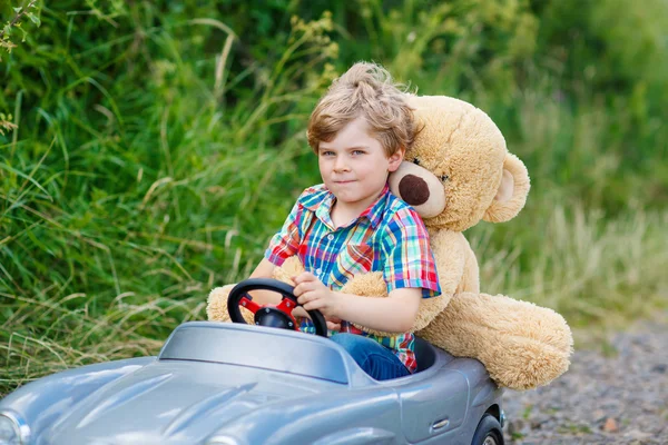 Niño pequeño conduciendo un coche de juguete grande con un oso, al aire libre . — Foto de Stock