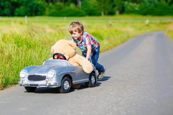 Kleiner Junge fährt großes Spielzeugauto mit einem Bären im Freien. — Stockfoto