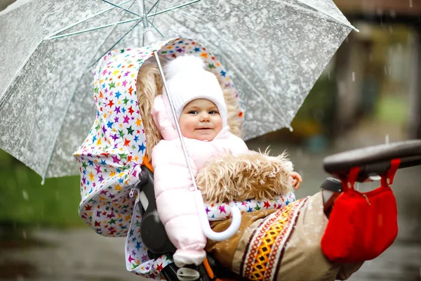 Niedliche kleine schöne Mädchen sitzen im Kinderwagen oder Kinderwagen an kalten Tagen mit Schneeregen, Regen und Schnee. Glücklich lächelndes Kind in warmer Kleidung, modisch stylischer Babymantel. Baby mit großem Regenschirm — Stockfoto