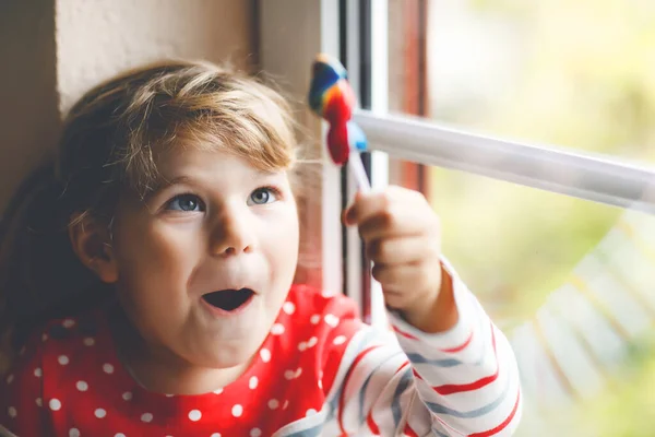 パンデミックコロナウイルスの隔離中に雨の日に窓のそばに座ってかわいい幼児の女の子。愛らしい子供食べるとなめる虹色のロリポップ — ストック写真