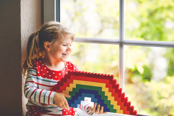 Roztomilé batole holčička po okně vytvořit duhu s barevnými plastovými bloky během pandemické koronavirus karantény. Děti vyrobeny a malovat duhy po celém světě jako znamení. — Stock fotografie