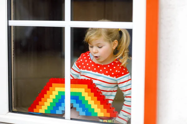窓によるかわいい幼児の女の子は、パンデミックコロナウイルスの隔離中にカラフルなプラスチックブロックを持つ虹を作成します。世界中の虹を看板として描く子どもたち. — ストック写真