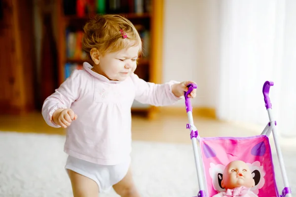 Schattig schattig baby meisje maken eerste stappen met pop rijtuig. Mooi peuter kind duwen kinderwagen met speelgoed thuis. Gelukkige dochter leren lopen en staan — Stockfoto