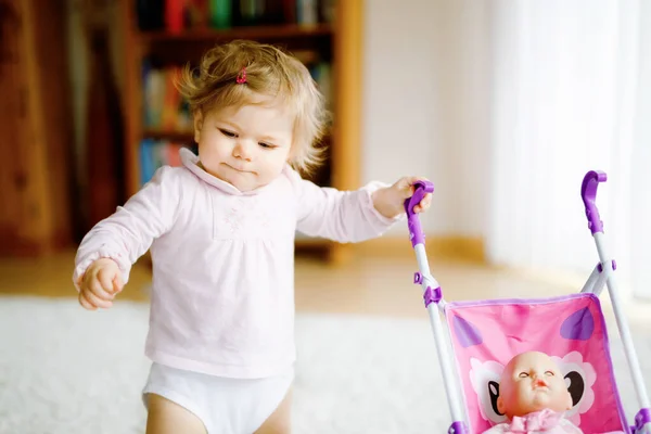 Linda menina adorável fazendo primeiros passos com carruagem de boneca. Criança bonita empurrando carrinho com brinquedo em casa. Filha feliz aprendendo andando e de pé — Fotografia de Stock