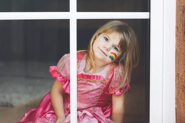 Niedliche einsame kleine Mädchen in Prinzessinnenkleid am Fenster sitzend mit Regenbogen mit bunten Farben im Gesicht während der Coronavirus-Quarantäne. Kinder basteln und bemalen Regenbogen auf der ganzen Welt — Stockfoto