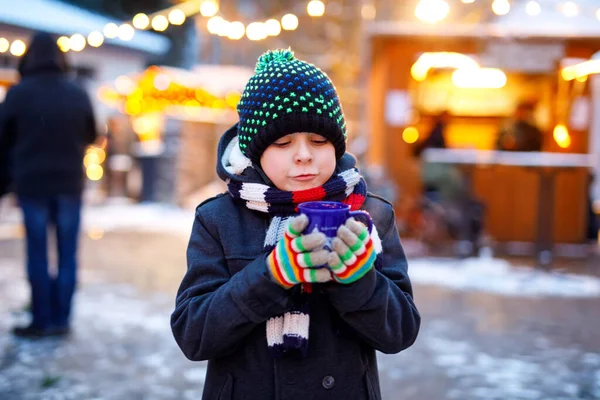 Niño lindo bebiendo ponche de niños calientes o chocolate en el mercado de Navidad alemán. Niño feliz en el mercado familiar tradicional en Alemania, niño risueño en ropa de invierno colorida — Foto de Stock