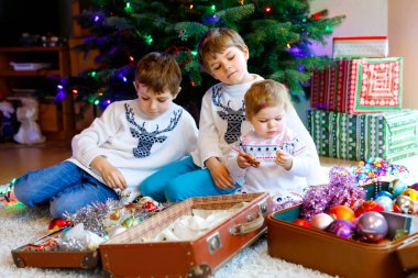 İki küçük oğlan ve sevimli kız bebek Noel ağacını eski model oyuncaklar ve toplarla süslüyorlar. Aile tatili için aile hazırlık kutlaması. Evde üç çocuk, erkek ve kız kardeş var..