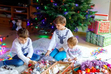 İki küçük oğlan ve sevimli kız bebek Noel ağacını eski model oyuncaklar ve toplarla süslüyorlar. Aile tatili için aile hazırlık kutlaması. Evde üç çocuk, erkek ve kız kardeş var..