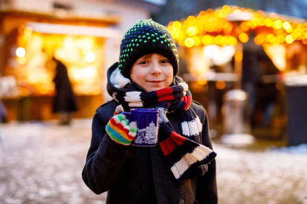Niño lindo bebiendo ponche de niños calientes o chocolate en el mercado de Navidad alemán. Niño feliz en el mercado familiar tradicional en Alemania, niño risueño en ropa de invierno colorida — Foto de Stock