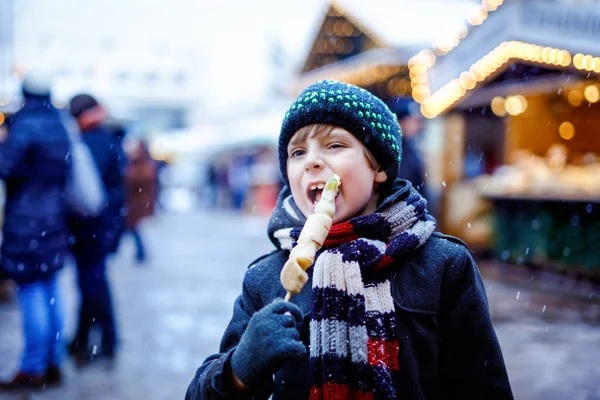 Little cute kid boy eating white chocolate covered fruits on skewer on traditional German Christmas market. Happy child on traditional family market in Germany during snowy day. — Stock Photo, Image