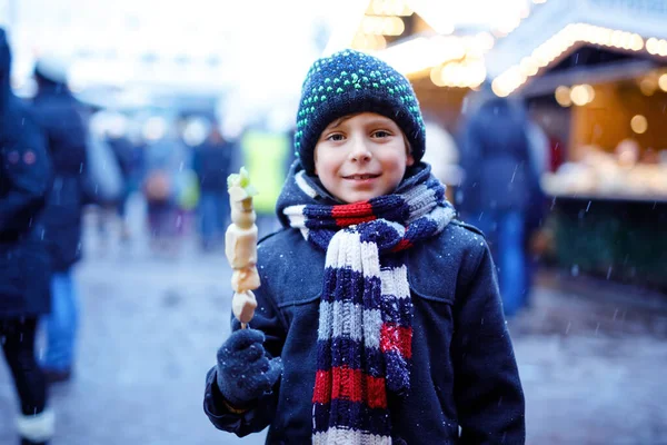 Der kleine süße Junge isst auf dem traditionellen deutschen Weihnachtsmarkt mit weißer Schokolade überzogene Früchte am Spieß. Glückliches Kind bei verschneitem Tag auf traditionellem Familienmarkt in Deutschland. — Stockfoto