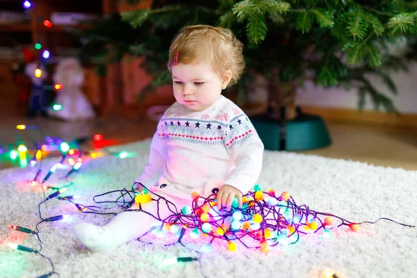 Entzückendes kleines Mädchen mit bunten Lichtergirlanden in niedlichen Händen. Kleines Kind in festlicher Kleidung schmückt Weihnachtsbaum mit Familie. Erstes Fest des traditionellen Feiertags namens Weihnachten — Stockfoto