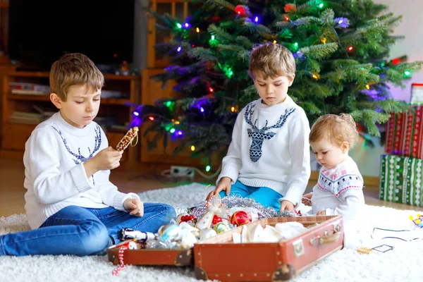 Zwei kleine Jungen und ein entzückendes Mädchen schmücken den Weihnachtsbaum mit altem Spielzeug und Bällen. Familienvorbereitungsfeier des Familienurlaubs. Drei Kinder, Geschwister zu Hause. — Stockfoto