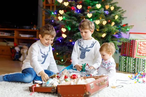 İki küçük oğlan ve sevimli kız bebek Noel ağacını eski model oyuncaklar ve toplarla süslüyorlar. Aile tatili için aile hazırlık kutlaması. Evde üç çocuk, erkek ve kız kardeş var.. — Stok fotoğraf