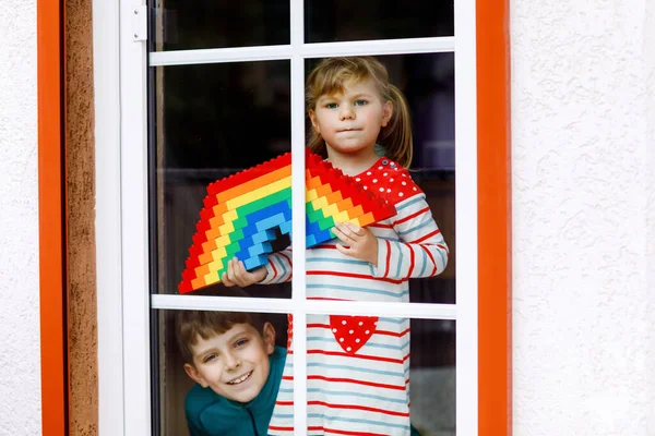 Küçük kız ve çocuk pencere kenarından pandemik koronavirüs karantinası sırasında renkli plastik bloklarla gökkuşağı oluşturuyorlar. İki çocuk, kardeşler dünyaya gökkuşağı çiziyor.. — Stok fotoğraf