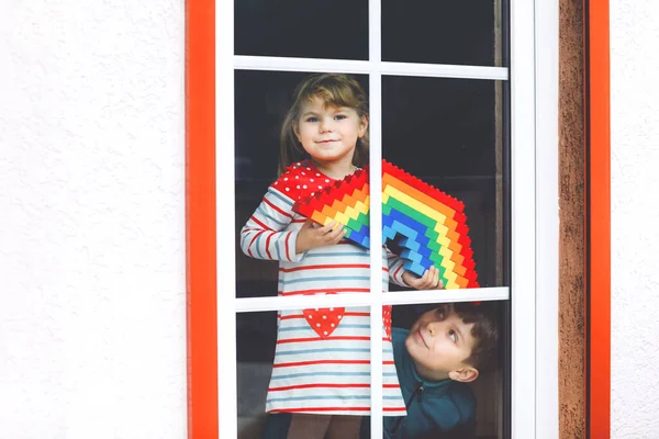 Småbarn flicka och pojke genom fönstret skapa regnbåge med färgglada plastblock under pandemisk coronavirus karantän. Två barn, syskon tillverkade och måla regnbågar runt om i världen som tecken. — Stockfoto