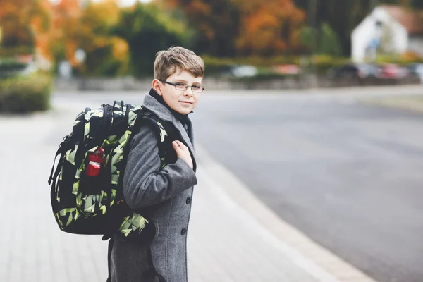 Glad pojke med glasögon och ryggsäck eller väska. Skolbarn i stilren fashon coan på väg till mellanstadiet eller gymnasiet på kall höstdag. Friska barn utomhus på gatan, på regnig dag. — Stockfoto